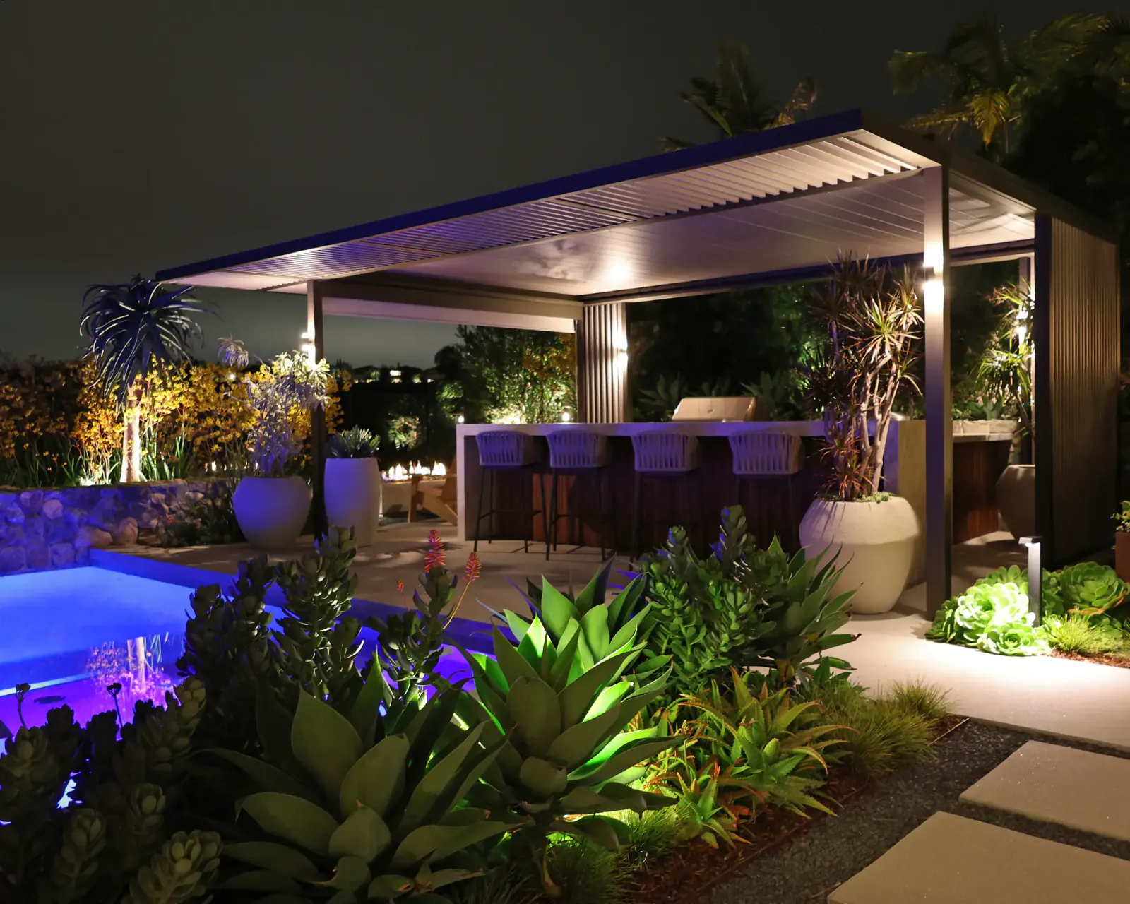 Uplit agaves and a lit louvered pergola over an outdoor bar beside a glowing pool at night